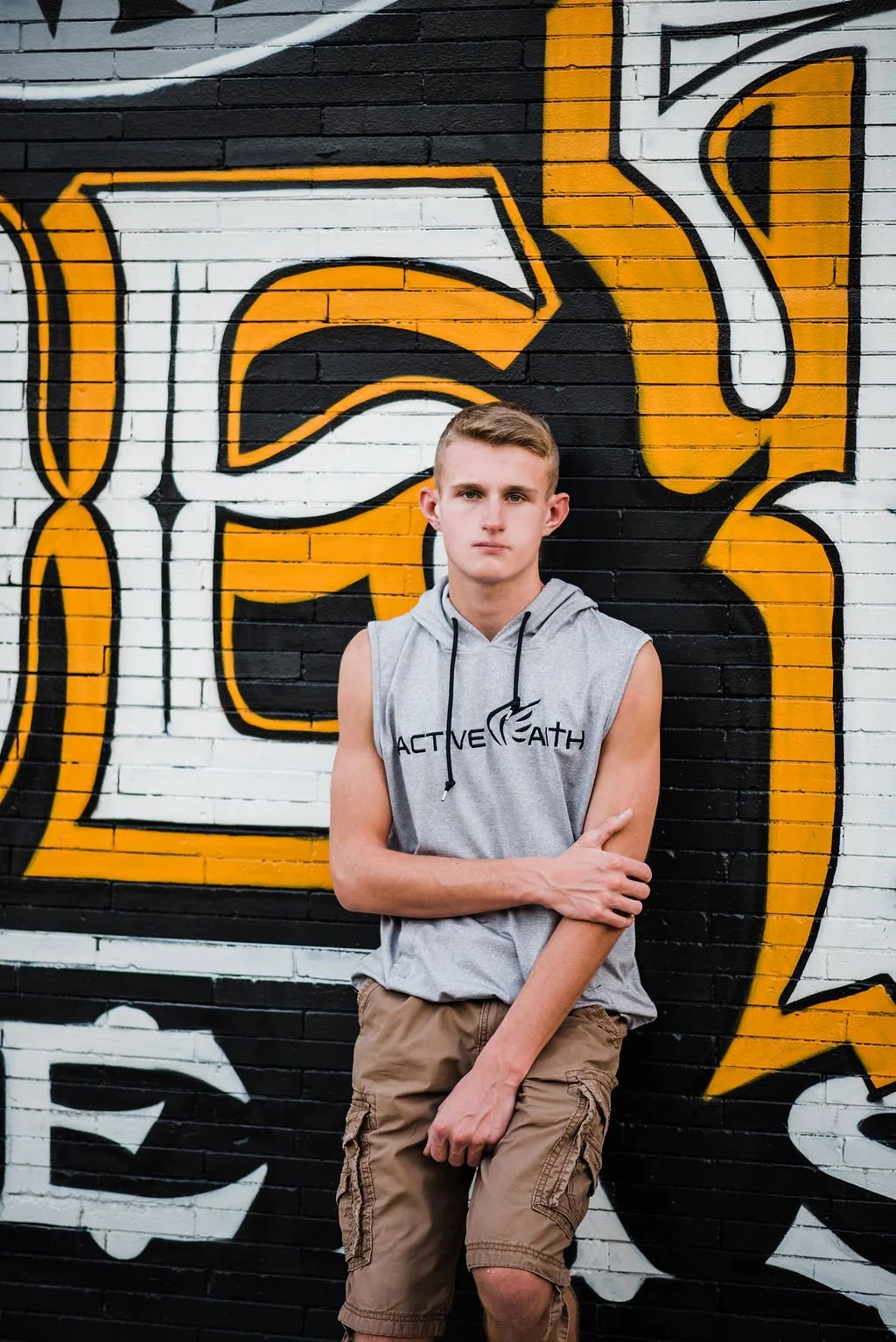 Young man with short hair wearing a sleeveless gray hoodie and cargo shorts stands in front of a graffiti wall with large black, white, and yellow letters.