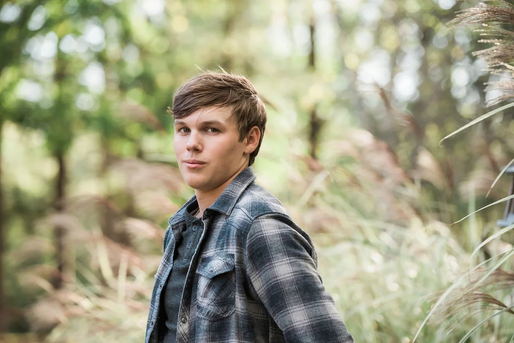 A young man with brown hair in a plaid shirt standing outdoors among green trees and tall grass.