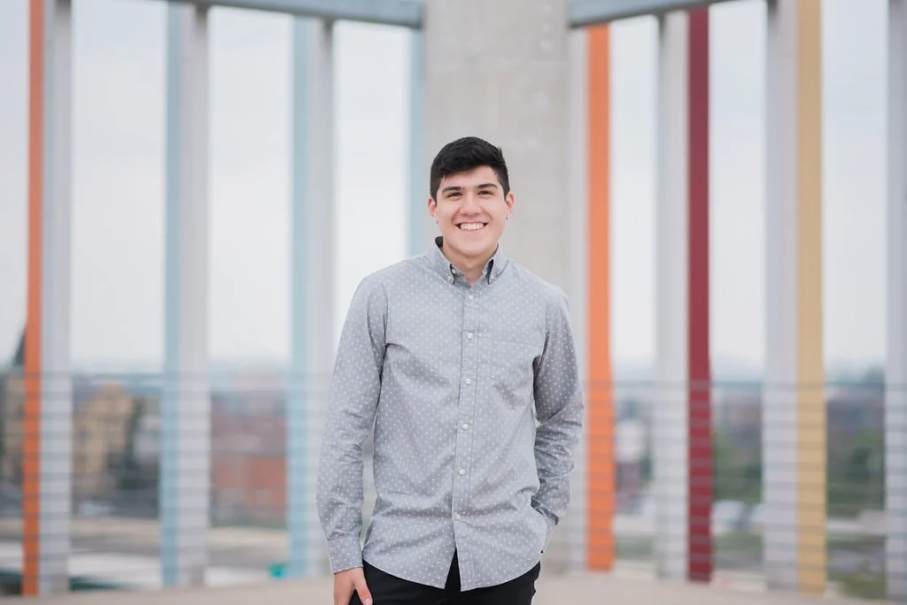 A young man with dark hair, wearing a light gray button-up shirt, smiling and standing outdoors in front of a modern building with colorful structural beams.