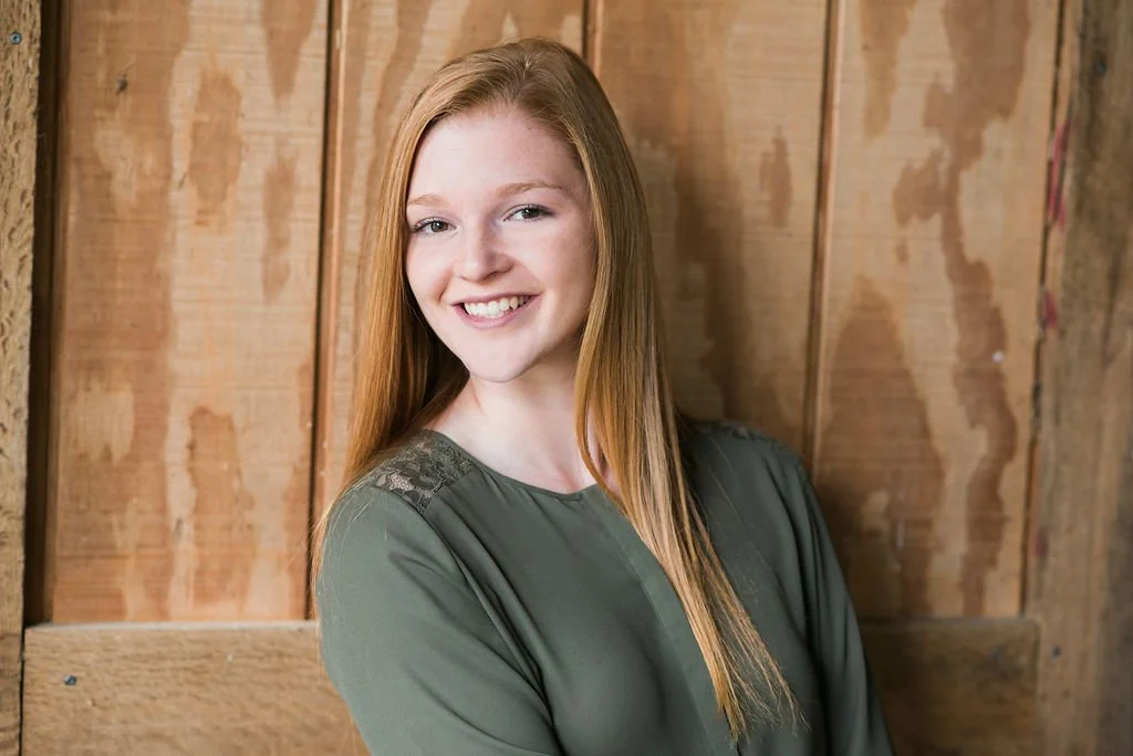 A young woman with long red hair smiling and leaning against a wooden wall.