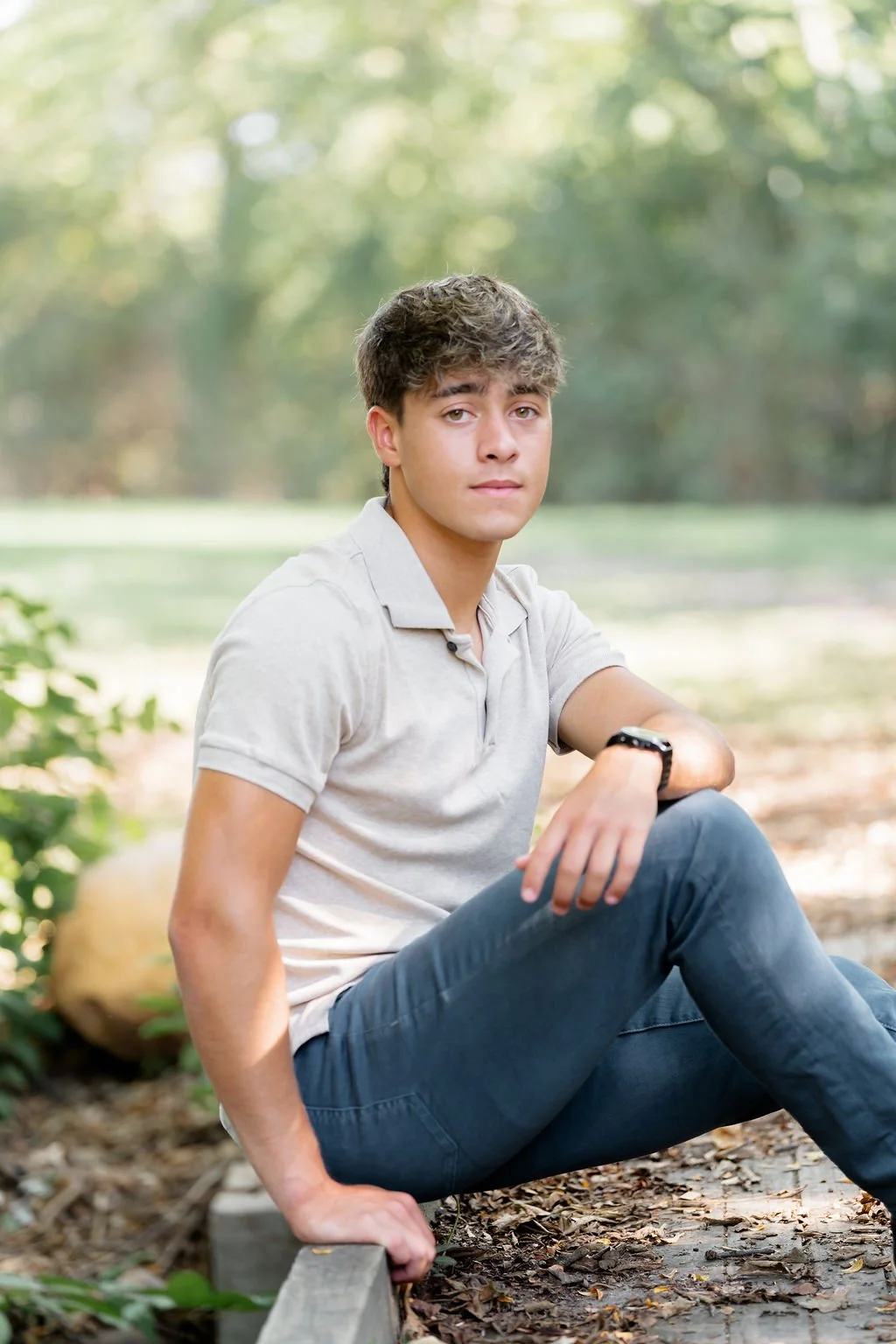 A young man with wavy brown hair sitting on a wooden curb in a park with trees and sunlight in the background, looking at the camera with a neutral expression.