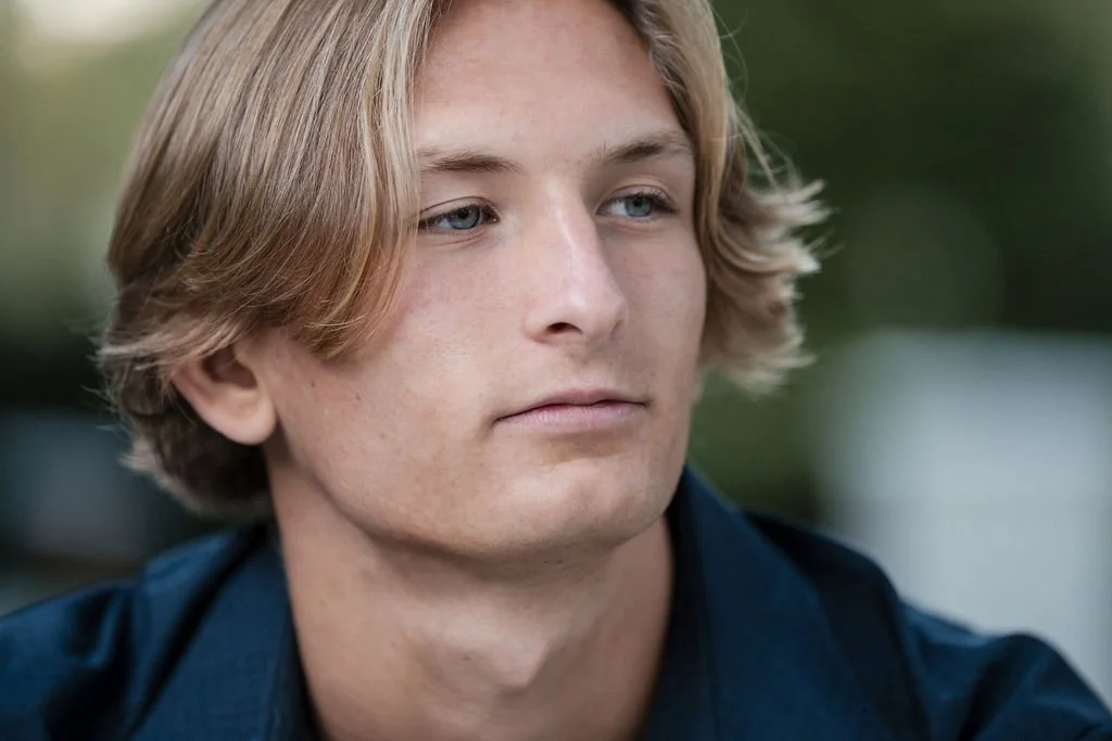 A young man with blonde hair and blue eyes looking to the side in an outdoor setting.