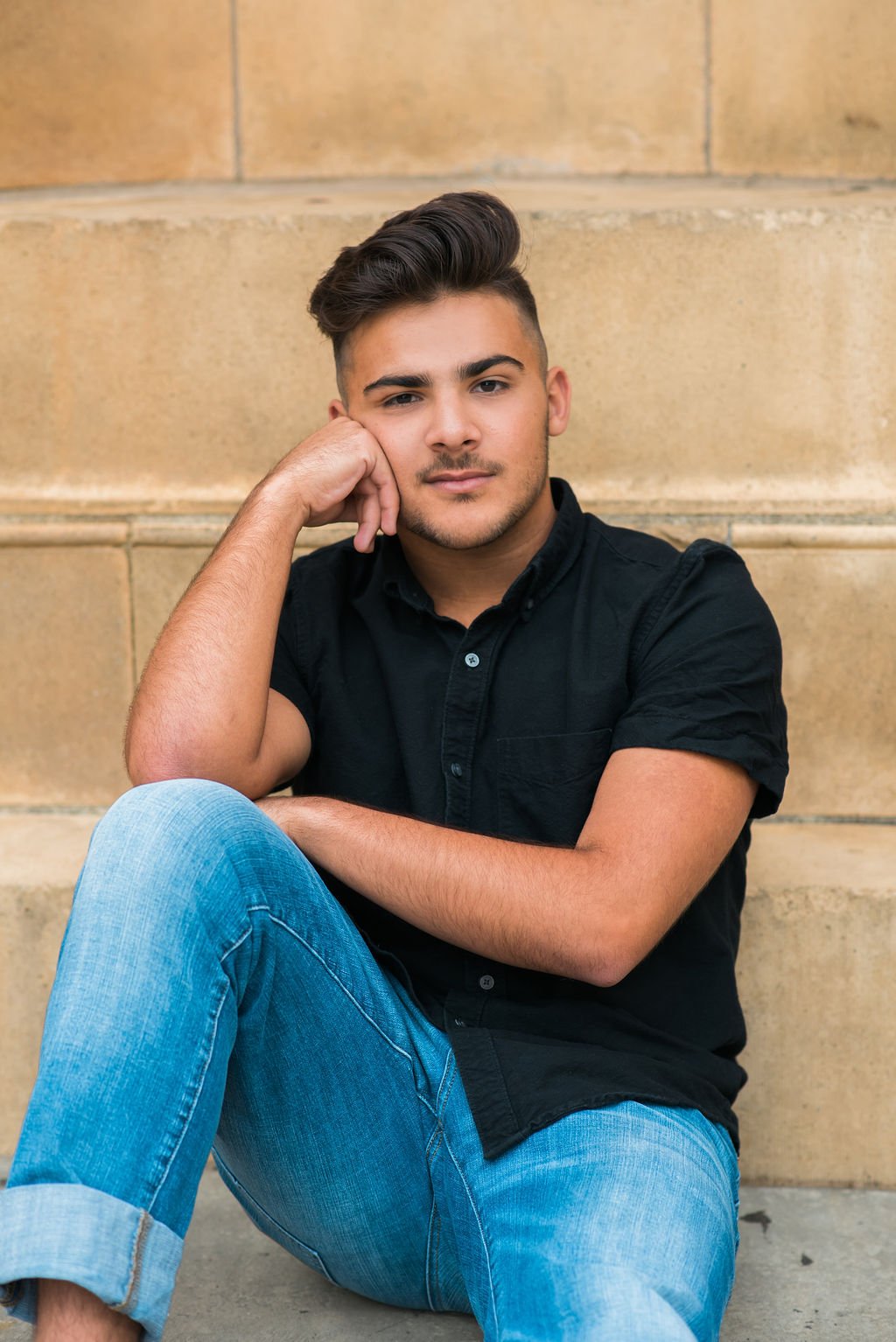 A young man with styled hair sitting against a stone wall, wearing a black short-sleeved button-up shirt and light blue jeans, looking at the camera with a relaxed expression.