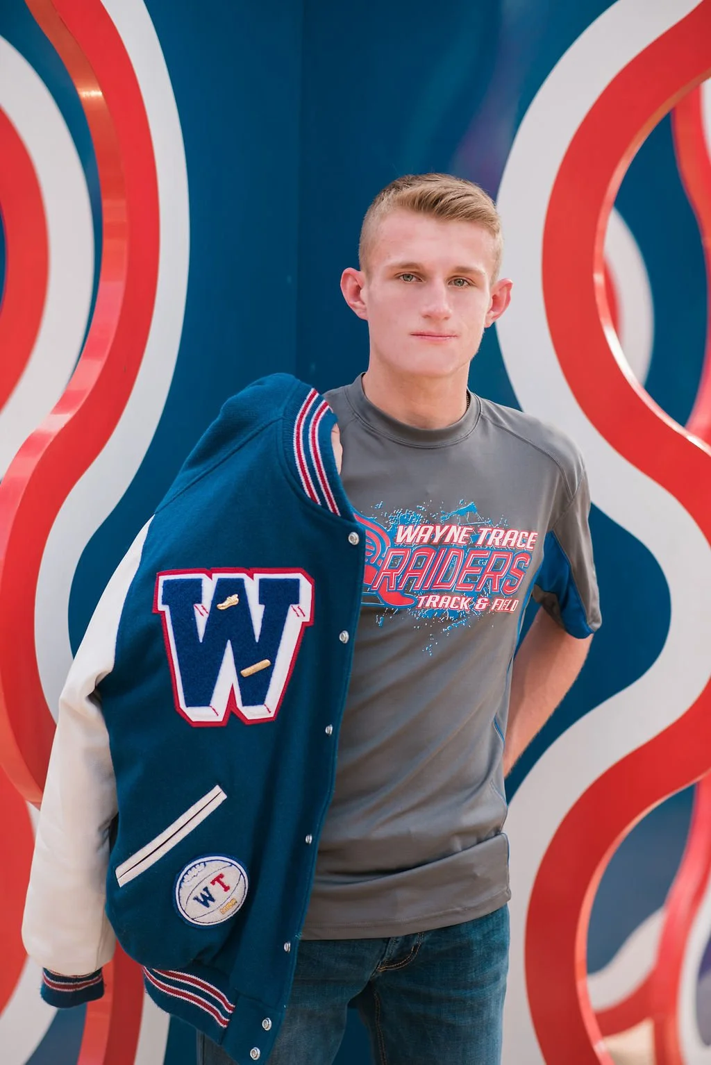 A young man wearing a gray Wayne Trace Raiders track and field T-shirt stands in front of a colorful, wavy background. He holds a blue letterman jacket with a large 'W' patch over his shoulder.