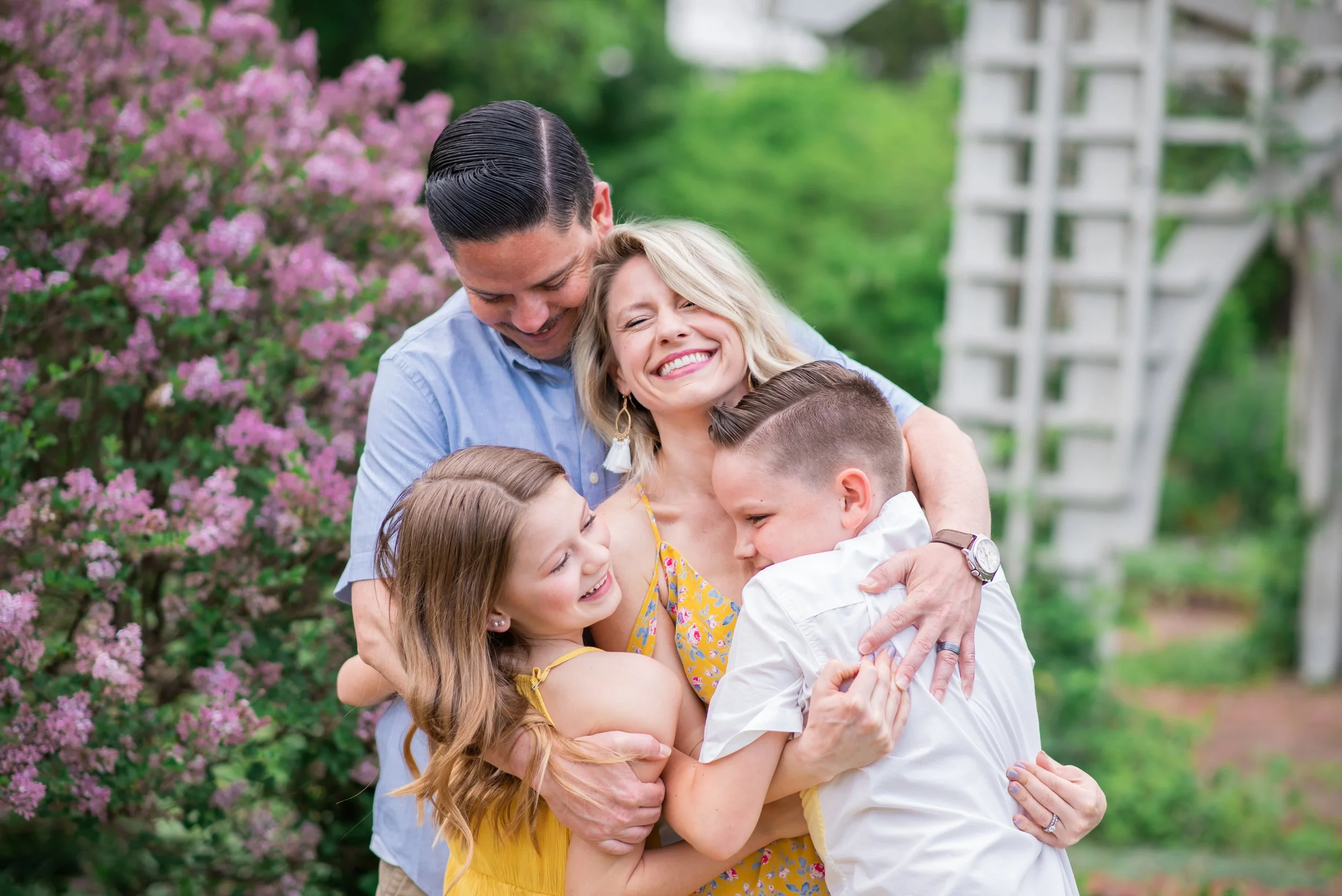 A family of five hugging and smiling in a garden with blooming pink flowers and greenery.