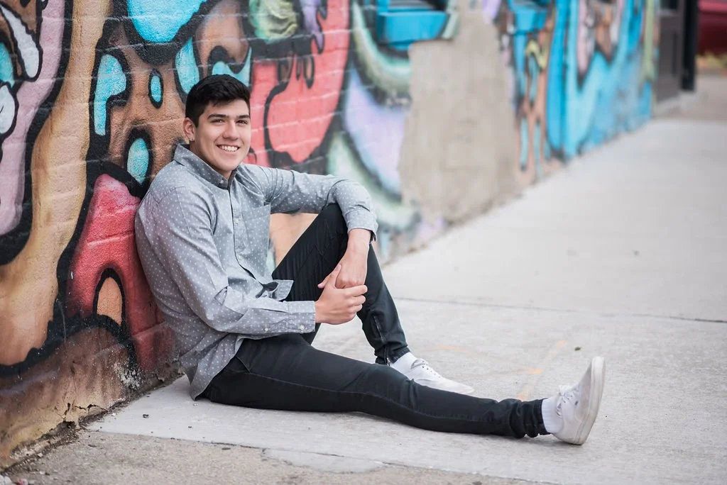A young man sitting on the sidewalk against a colorful graffiti wall, smiling at the camera.