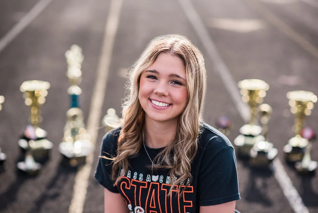 A young woman with long, wavy blonde hair smiling while sitting on an outdoor track field with multiple trophies behind her.