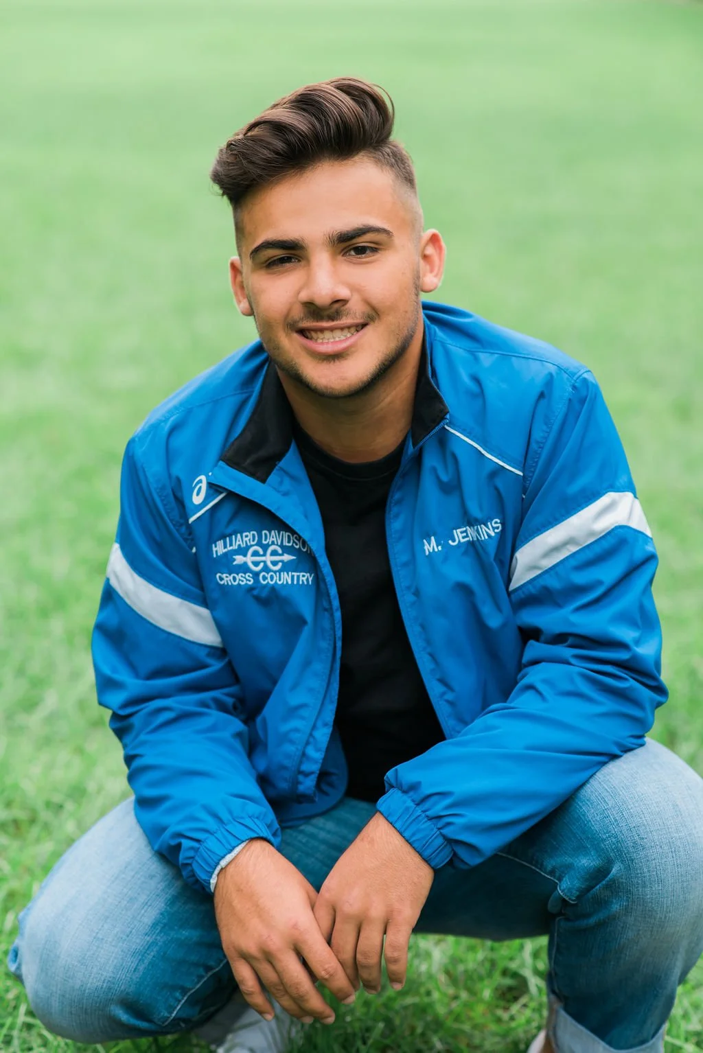 A young man with styled dark hair and light skin smiling while crouching on grass in a park. He is wearing a blue track jacket with white and black accents and the words "Hilliard Davidson Cross Country" and 