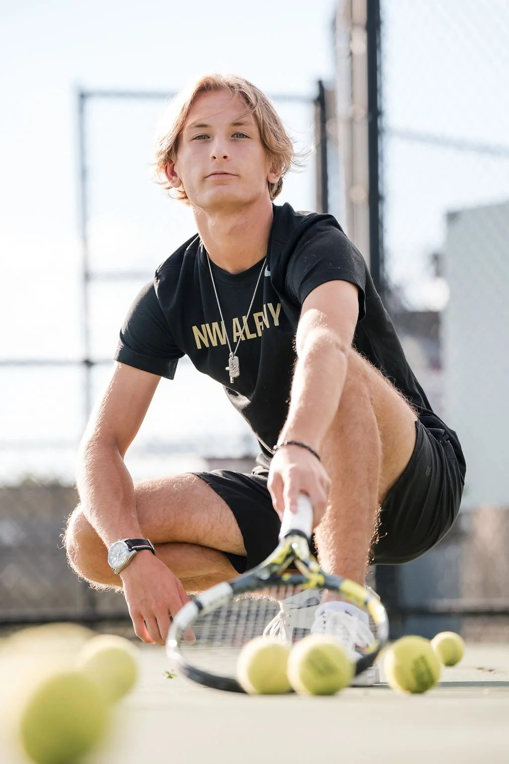 Young man crouching on tennis court, holding tennis racket surrounded by tennis balls, wearing black t-shirt, black shorts, wristwatch, and necklace.
