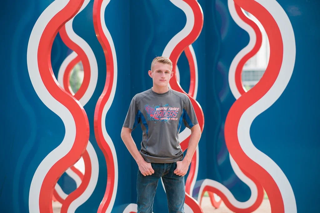 A young man stands in front of an abstract blue and red wavy art installation, wearing a gray sports T-shirt with 'Wayne Trace Raiders Track & Field' logo, with his hands in his pockets and looking at the camera.