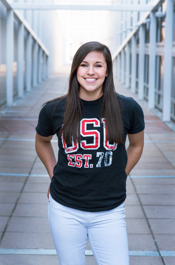 Young woman smiling, standing on a bridge or walkway, wearing a black T-shirt with red and white "USC EST. 70" written on it, and white pants.
