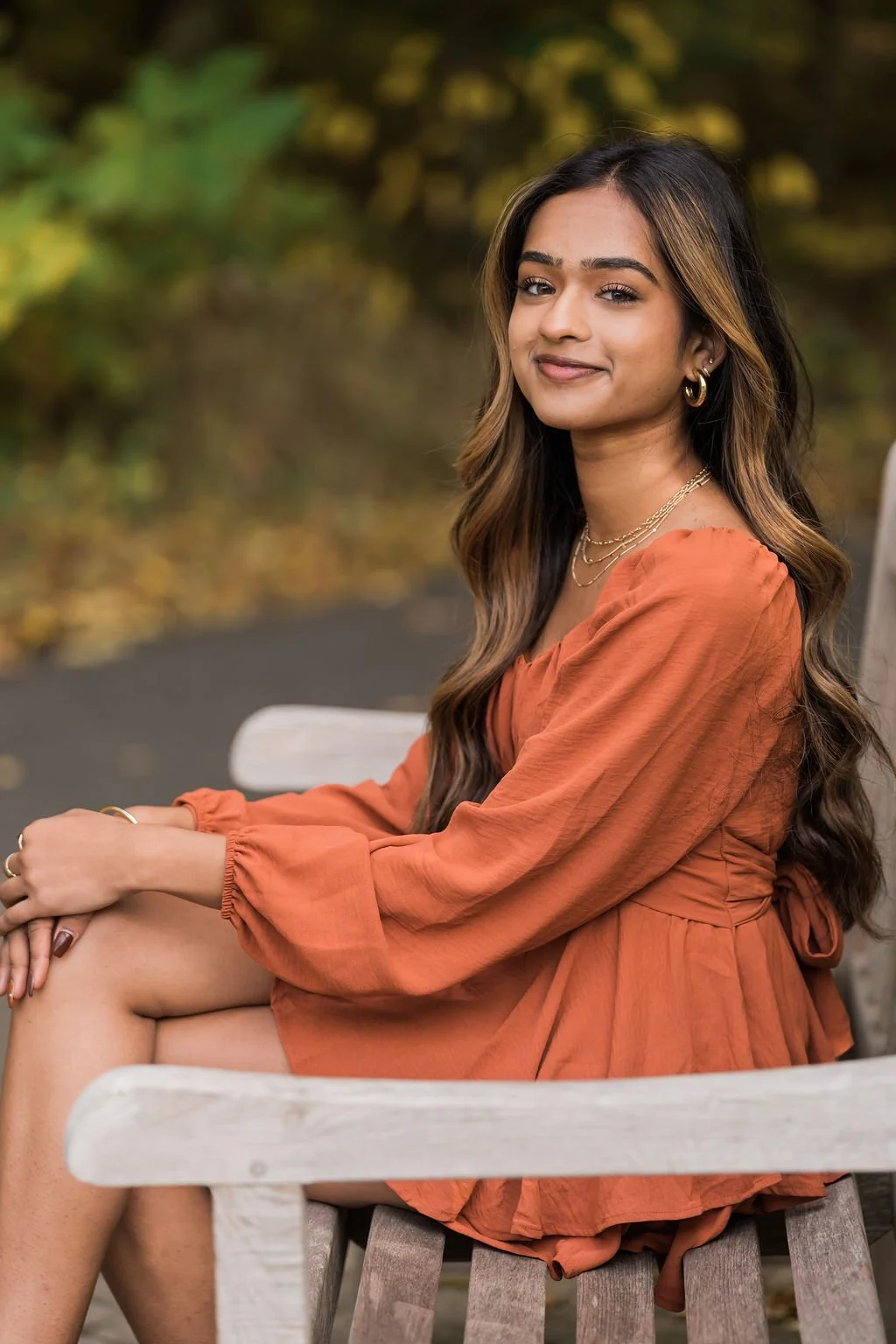 A young woman sitting on a wooden bench outdoors, wearing an orange dress with long sleeves, gold jewelry, and wavy hair, with a blurred background of green and brown foliage.