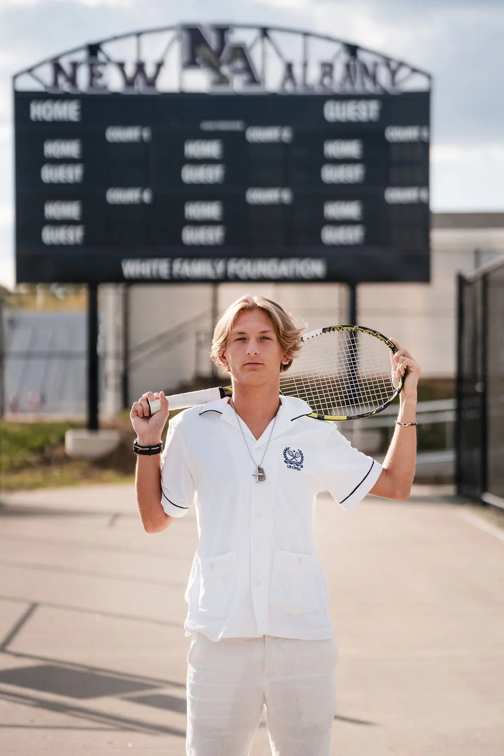 A young person standing on a tennis court holding a tennis racket over their shoulder. They are wearing a white polo shirt with a logo and a necklace, with a scoreboard in the background displaying 'Home' and 'Guest'.