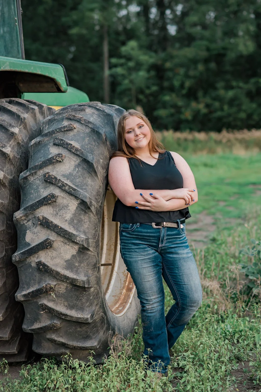 Smiling young woman with light skin and blond hair leaning against the large tire of a green tractor in a field with green grass and trees in the background.