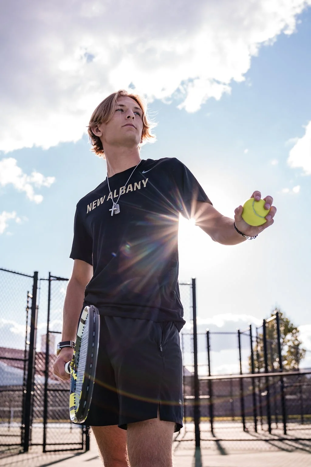 A young male tennis player standing outdoors on a tennis court holding a tennis ball in his right hand and a racket in his left, with the sun shining behind him.