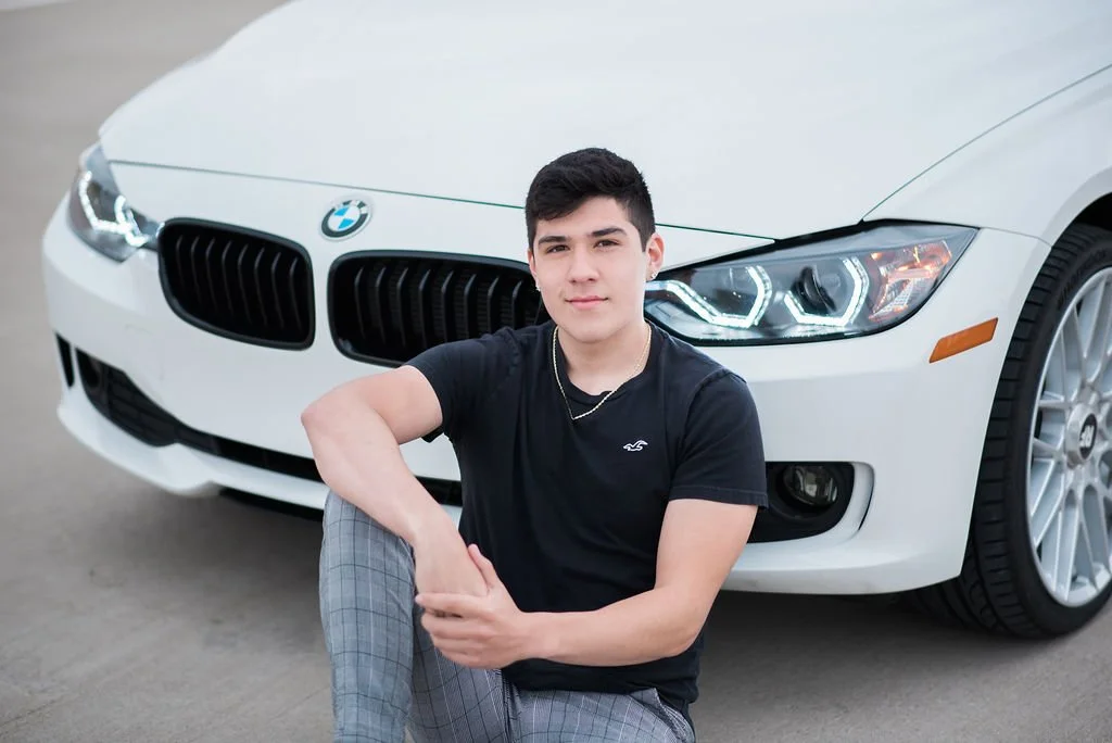 A young man sitting in front of a white BMW car with the hood closed, on a gray concrete surface.