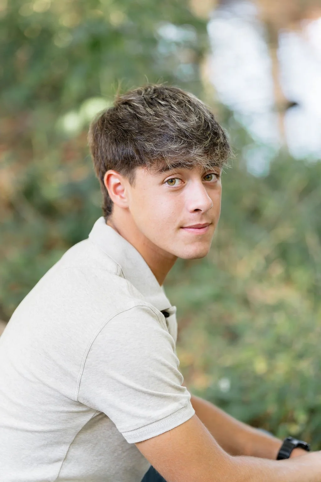A young man with light brown hair and hazel eyes looking over his shoulder outdoors, wearing a light-colored short-sleeve shirt, with blurred trees in the background.