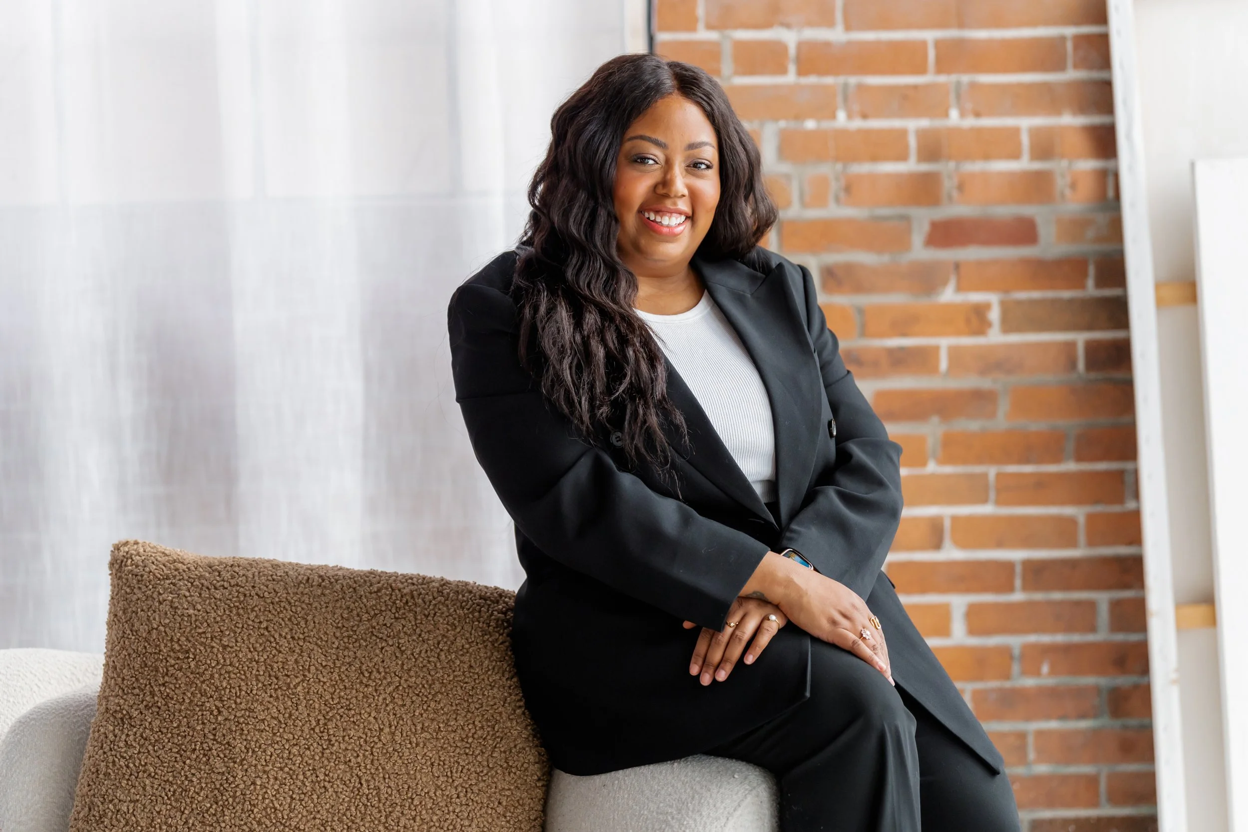 A woman with long curly black hair smiling and sitting on a beige couch with a brown cushion, wearing a black blazer and white top, against a backdrop of a brick wall and white curtains.