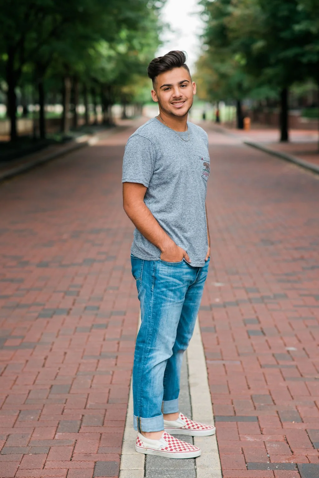 A young man stands on a brick-paved street lined with trees, wearing a gray t-shirt, blue jeans, and checkered slip-on shoes, smiling at the camera.