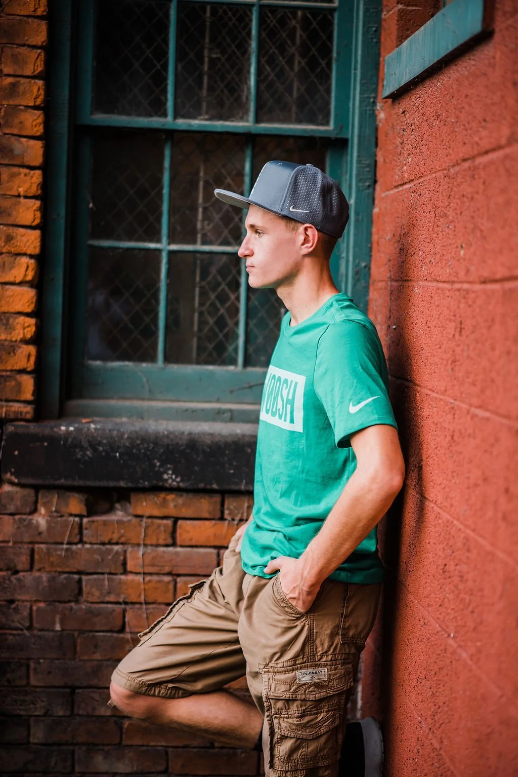A young man leaning against a red brick wall next to a green window with metal bars, in an urban setting. He is wearing a gray and black baseball cap, a green T-shirt with the word 'CROSH' on it, and tan cargo shorts, with his hands in his pockets an