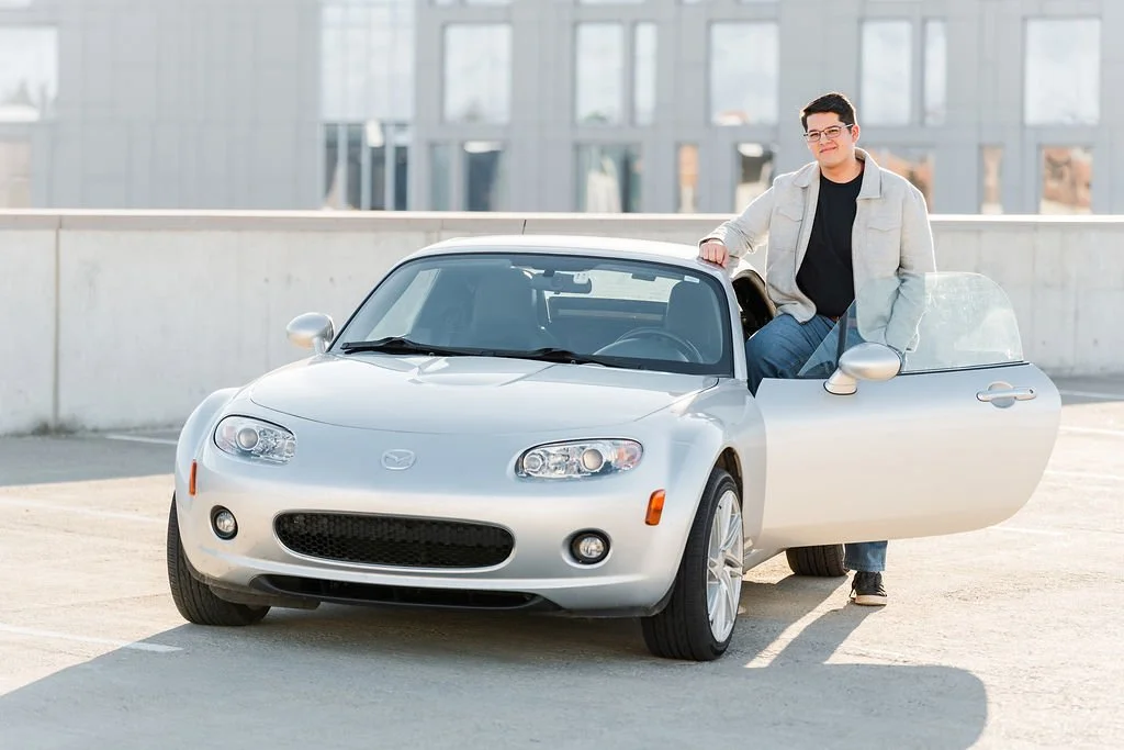 A young man with glasses, wearing a beige jacket and black shirt, standing next to a silver Mazda convertible in a parking lot.