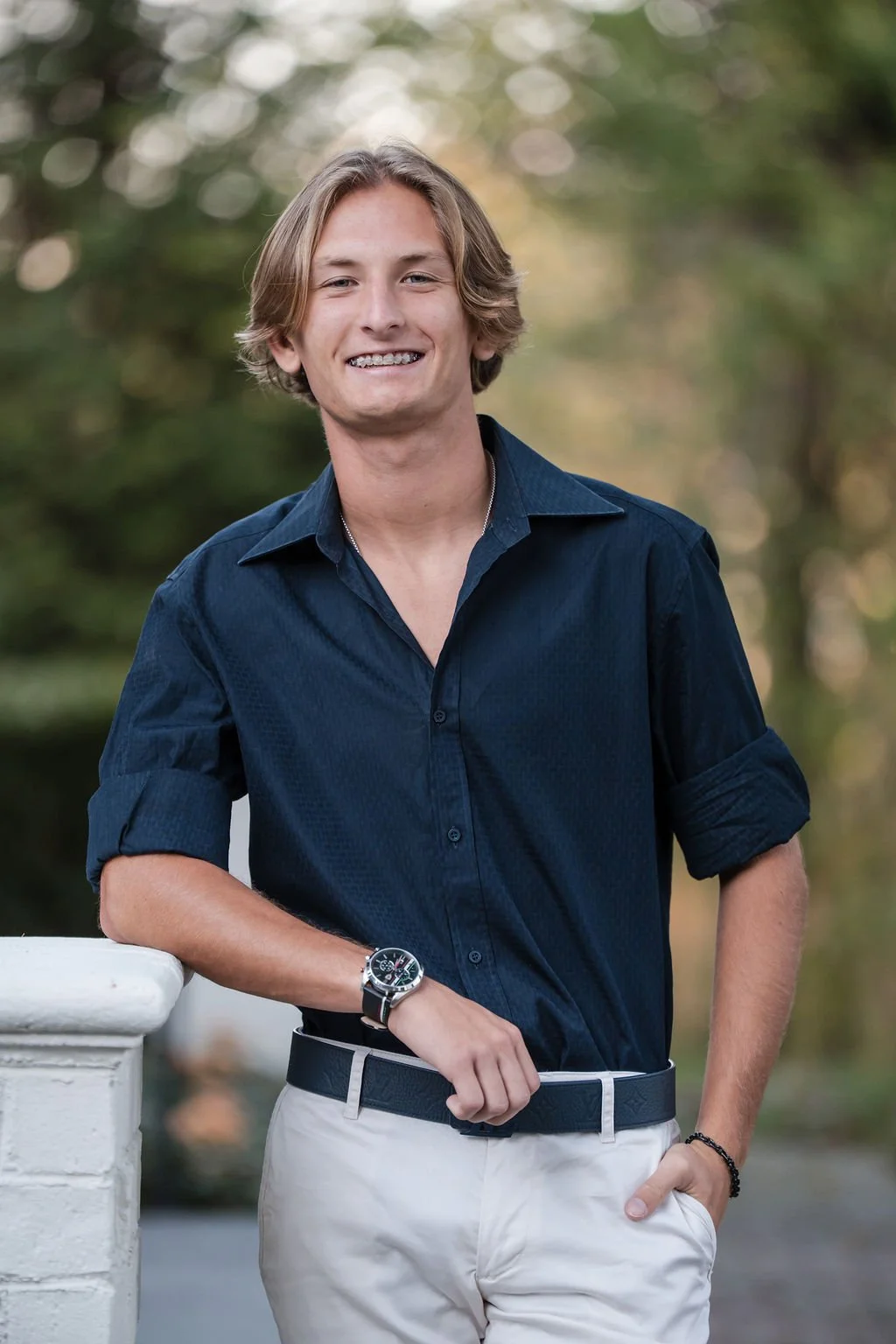 Young man with blonde hair wearing a navy blue shirt and beige pants, standing outdoors in front of blurred green foliage, smiling with braces.