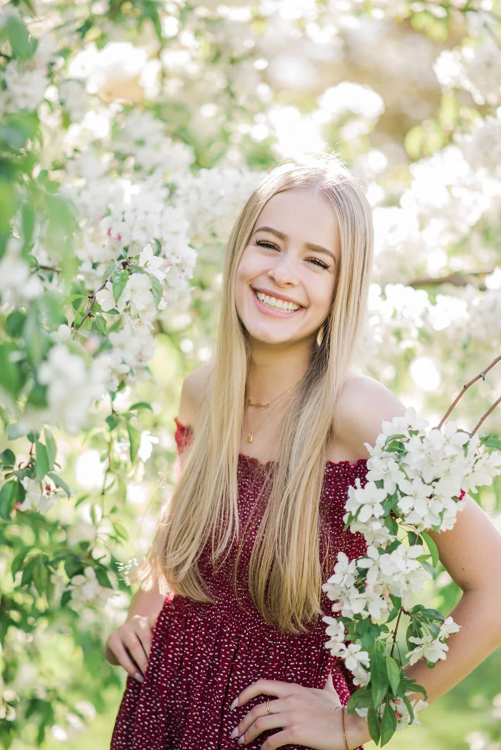 A smiling young woman with long blonde hair and light skin, wearing a red dress, standing among white flowering branches outdoors with sunlight filtering through the trees.