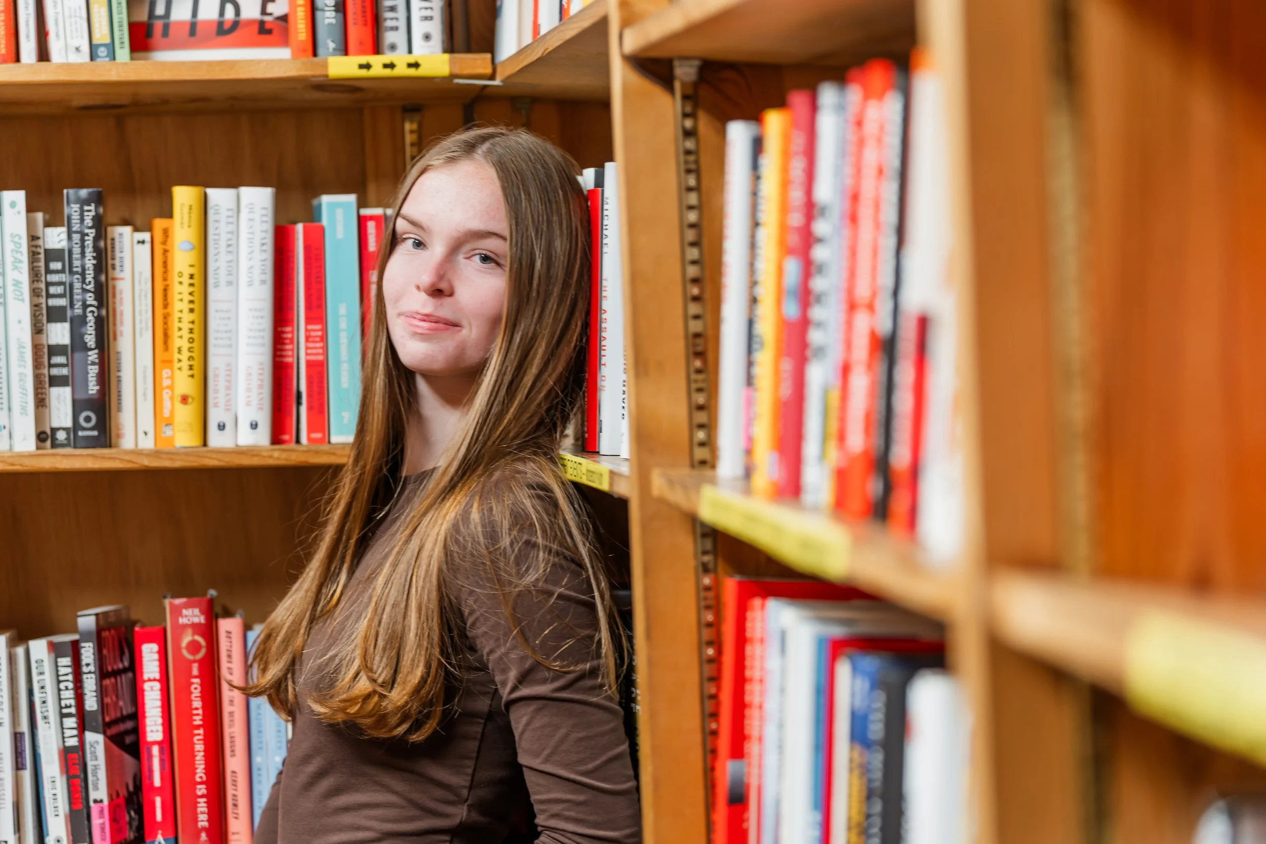 A young woman with long brown hair stands between bookshelves in a library, looking at the camera with a slight smile.