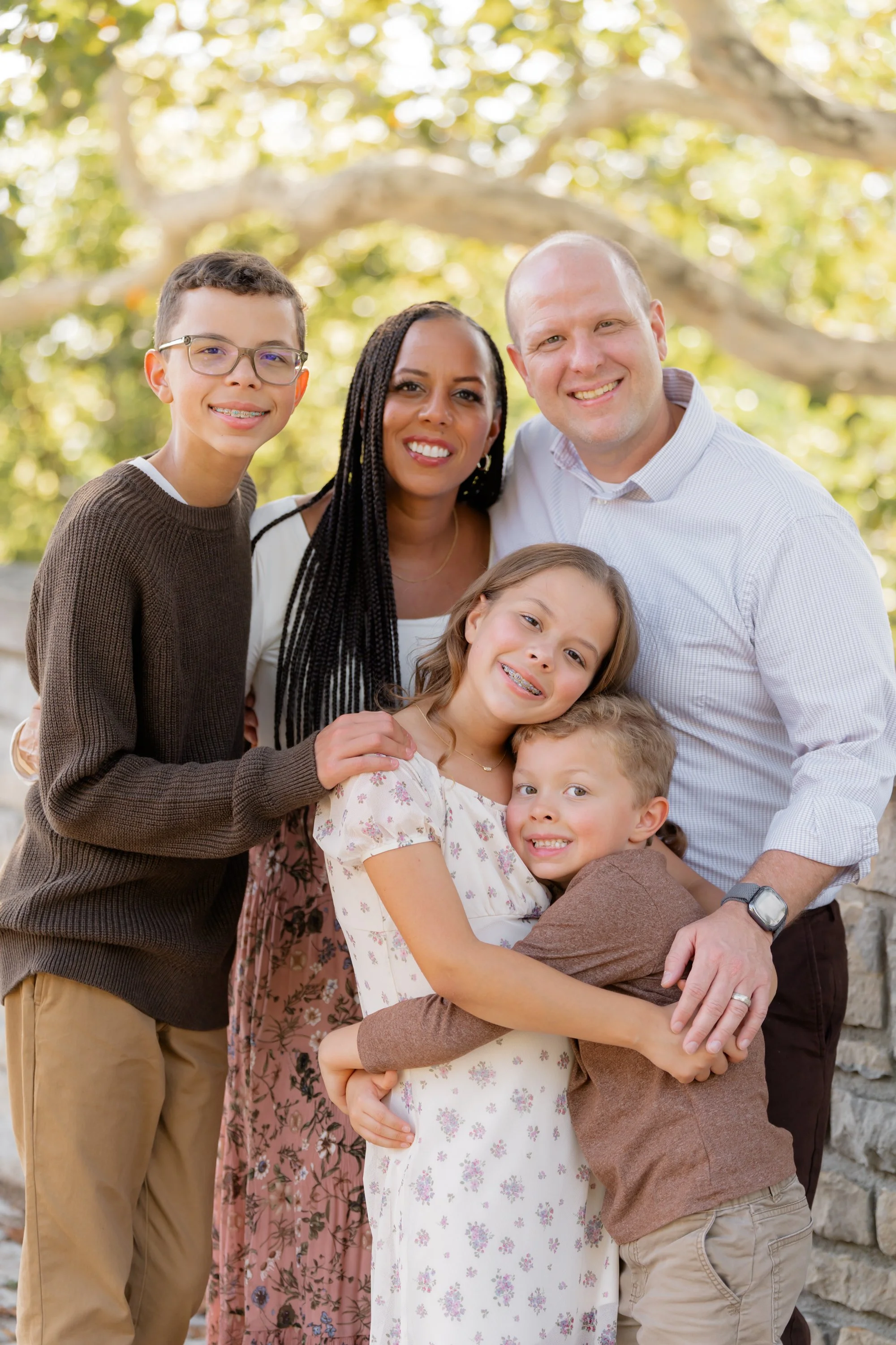 A family of six smiling and hugging outdoors near a stone wall, with trees and sunlight in the background.