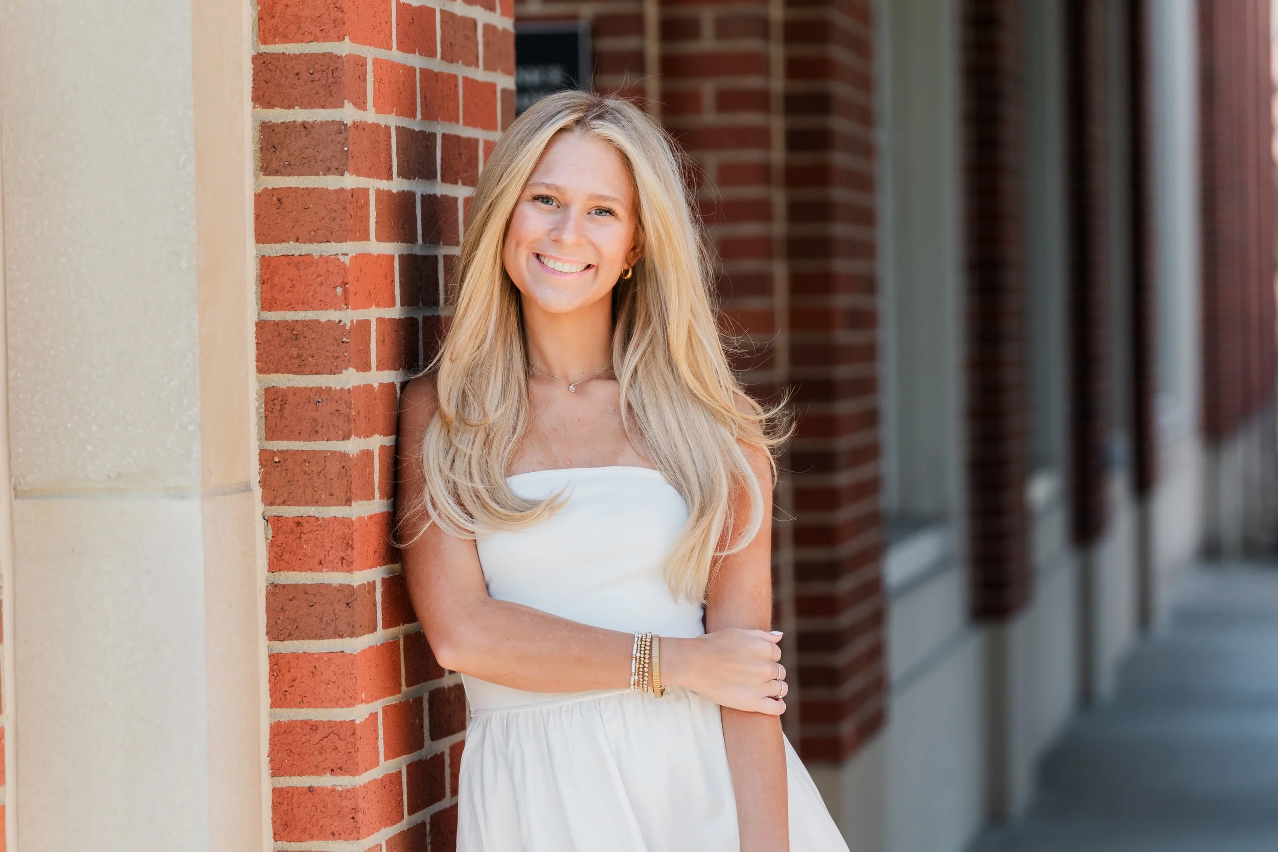 A young woman with long blonde hair, smiling, leaning against a red brick wall outdoors on a sunny day. She is wearing a white strapless dress and gold bracelets.