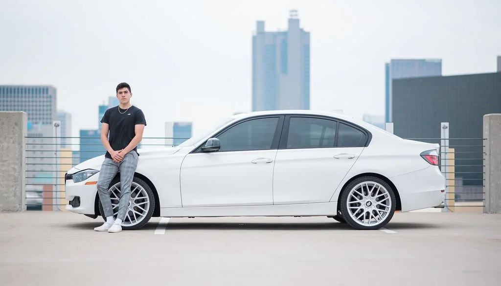 A young man standing next to a white sedan car on a rooftop parking lot with a city skyline in the background.