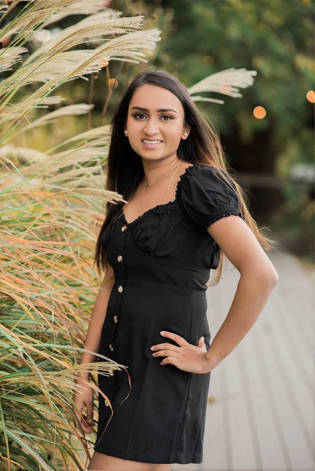 A young woman with long dark hair wearing a black dress with puffed sleeves and gold buttons, standing outdoors next to tall ornamental grass, with a blurred natural background.