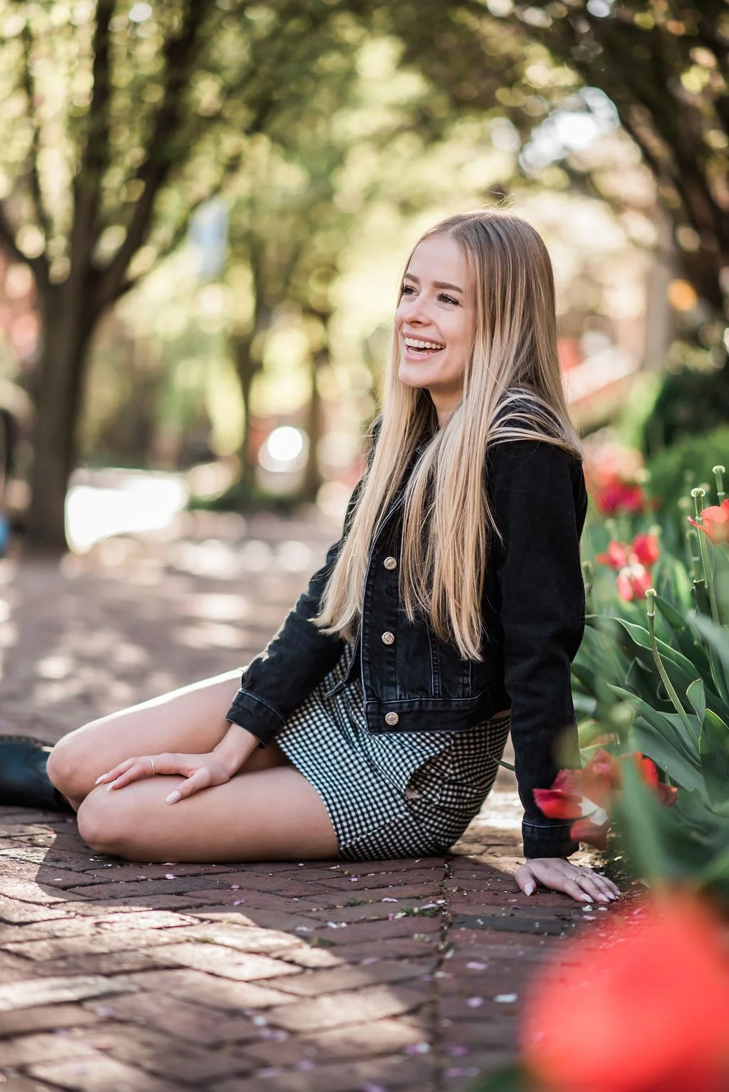 A young woman with long blonde hair sitting on a brick pathway outdoors, smiling and enjoying a sunny day, surrounded by green trees and pink flowers.