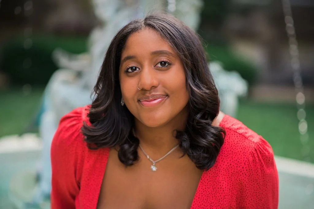 A young woman with dark, shoulder-length hair styled in loose waves, wearing a red top and a silver necklace with a small pendant, smiling softly outdoors with a fountain and greenery in the background.