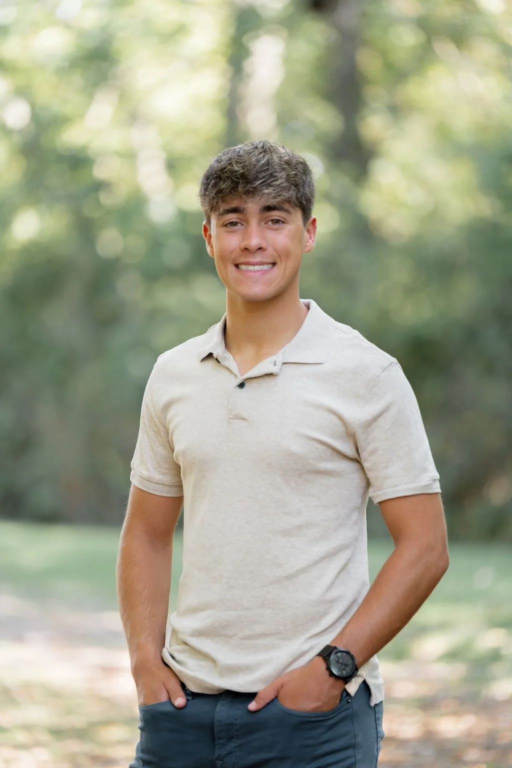 A young man smiling outdoors in a park or forest setting, wearing a beige polo shirt, dark pants, and a black wristwatch.