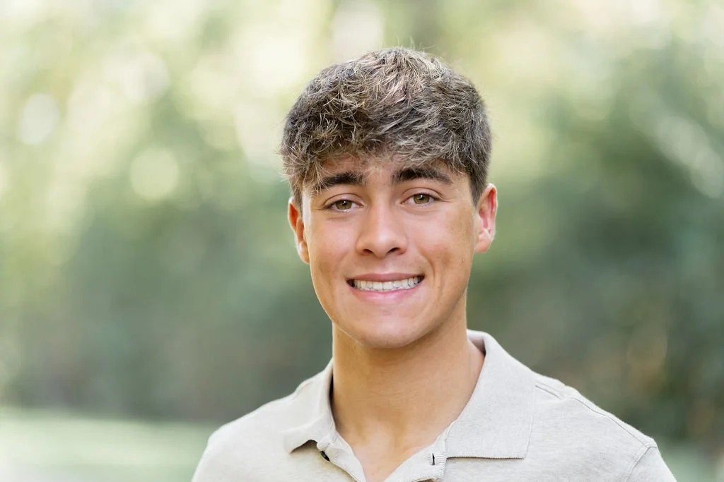 A young man with curly brown hair smiling outdoors in a park.