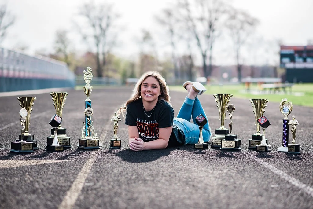 Young girl lying on a track field surrounded by various trophies.