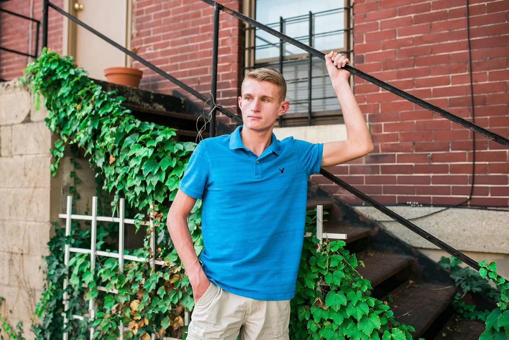A young man in a blue polo shirt and beige shorts standing outdoors beside a staircase, leaning on a black railing with one hand, with green ivy and a brick building in the background.