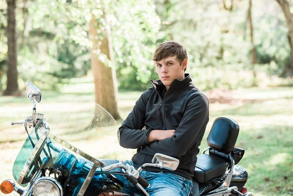 A teenage boy sitting on a motorcycle in a wooded outdoor park, wearing a black jacket with arms crossed and looking serious.