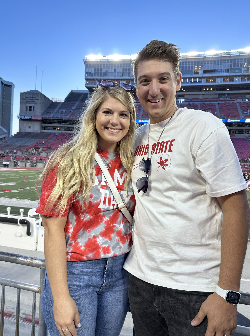A young couple smiling at the camera stands in a stadium after a football game, with empty red seats and a large scoreboard visible in the background.