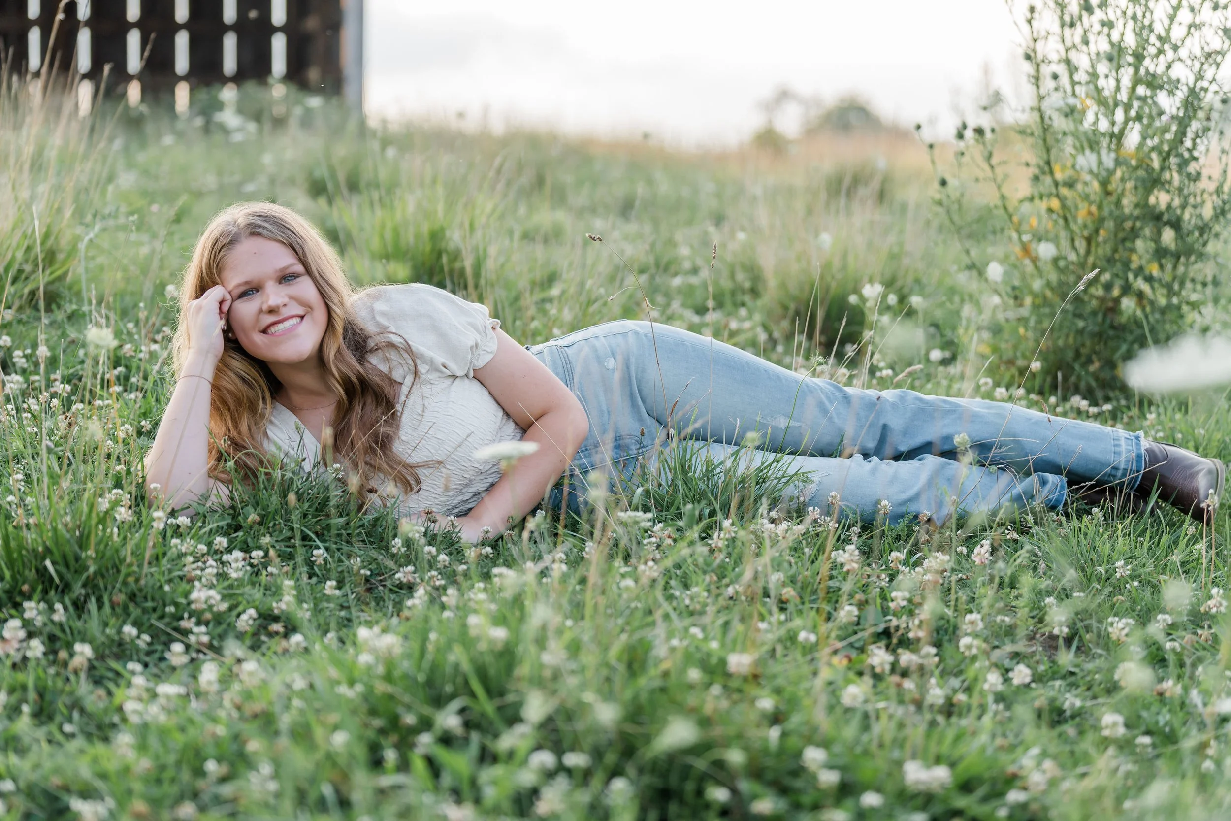 A woman with long, wavy hair lying on her side in a grassy field, smiling at the camera.