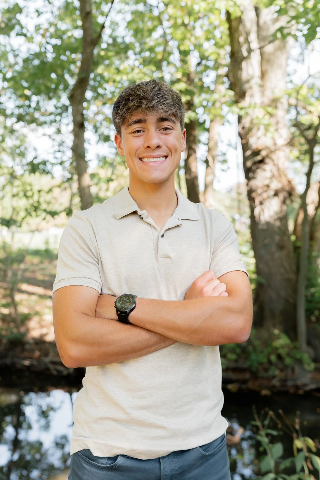 A young man with brown hair smiling and crossing his arms outdoors in a wooded area with trees and sunlight filtering through the leaves.