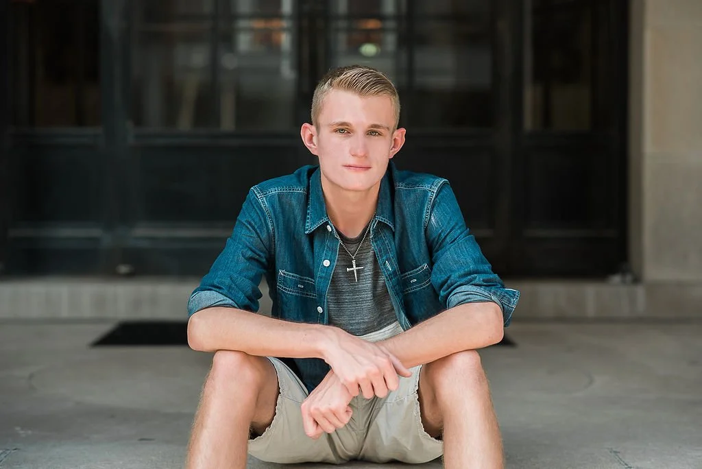 Young man sitting on concrete outside a building, wearing a denim shirt and shorts, with a cross necklace, looking at the camera.