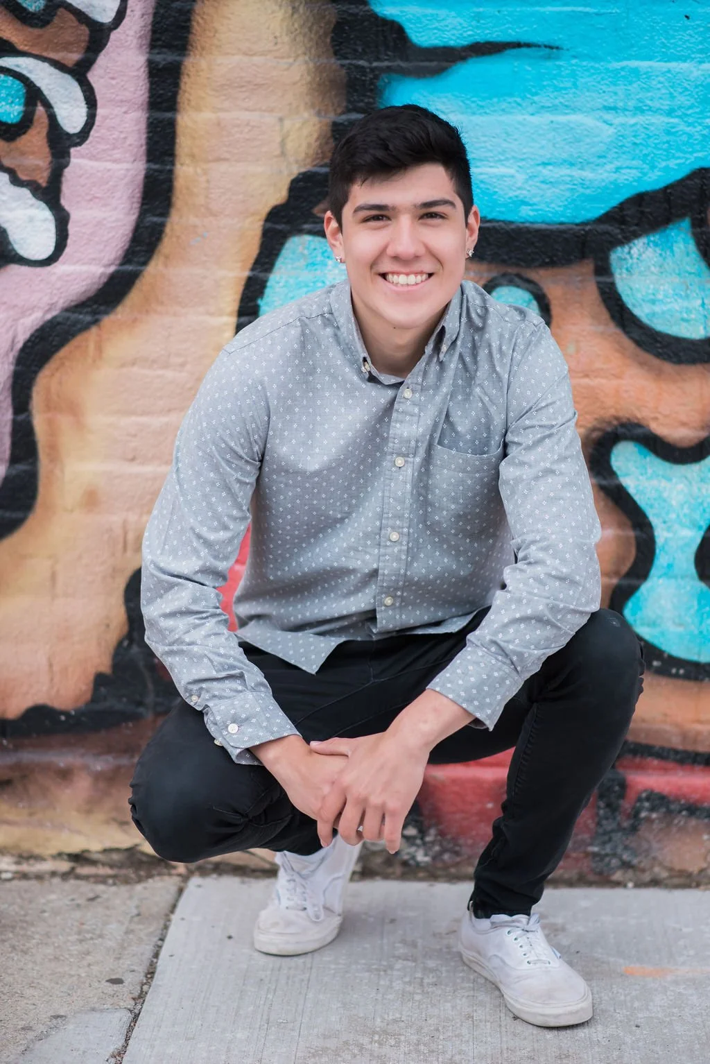 A young man with short dark hair, wearing a light gray dotted button-up shirt, black pants, and white sneakers, crouching in front of a colorful graffiti wall.