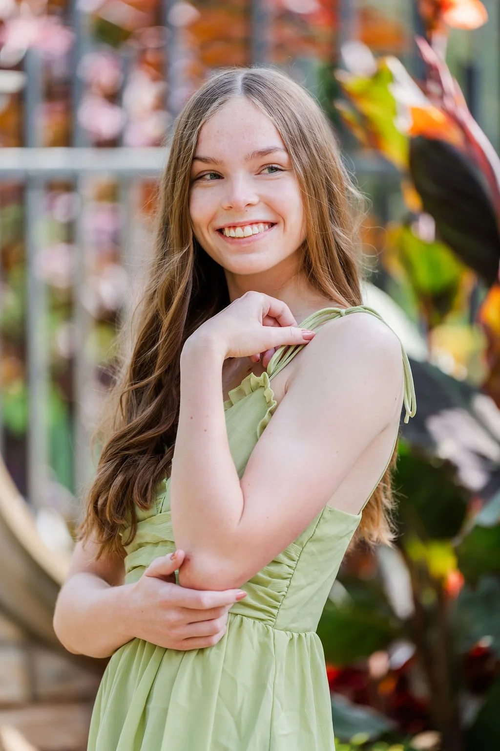 A young woman with long, wavy brown hair and fair skin, smiling and posing outdoors in front of a background of colorful plants and flowers. She is wearing a light green sleeveless dress and has her right hand touching her shoulder.