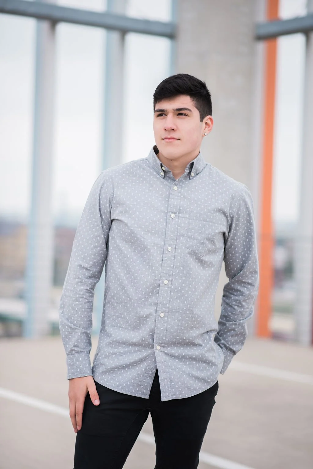 A young man with short dark hair wearing a gray button-up shirt with small white polka dots and black pants, standing outdoors near a modern building with glass and metal structures.