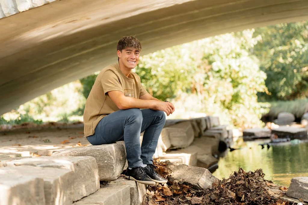 Young man sitting by a creek under a bridge, smiling at the camera.