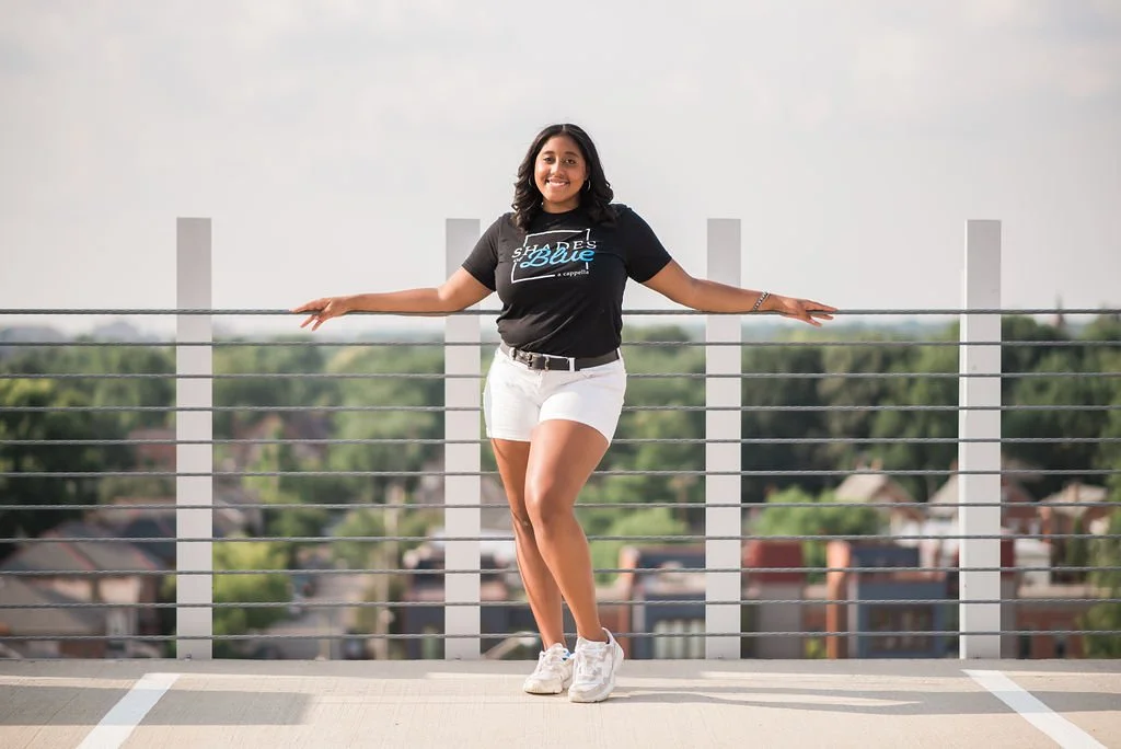 A young woman with dark hair and a bright smile, wearing a black T-shirt and white shorts, stands on a rooftop with a cityscape in the background, holding onto the rooftop railing.