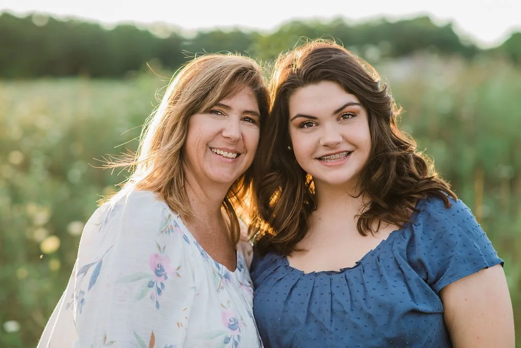 A mother and daughter posing outdoors together during sunset, both smiling at the camera, with a blurred green landscape background.
