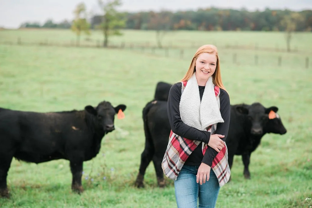 A young woman standing in a grassy field with three black cows behind her.